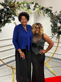 Two guests in evening attire posing beneath a floral arch on the red carpet